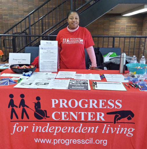 image of woman standing behind Progress Center resource table. Table has red covering and information packets are laid out across the table
