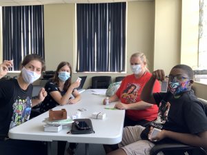 Image of four people, sitting at table. People are all wearing Covid 19 Pandemic Masks, and are all looking at camera. 