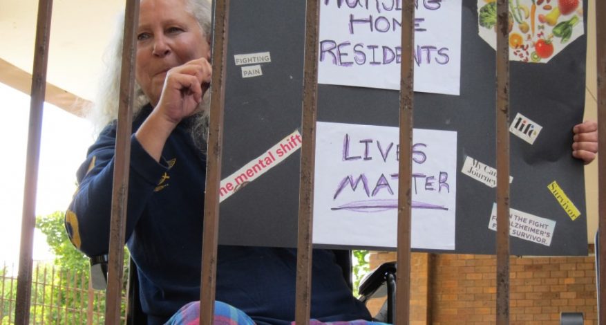 A woman in a wheelchair, in the court yard of a nursing home, behind a fence with tall iron bars. Woman is holding a sign that says Nursing Home Residents and a sign that says Lives Matter